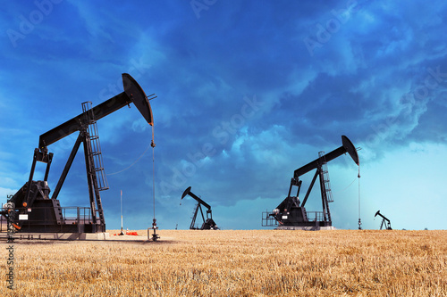 Four traditional pump jacks stand silhouetted against a dramatic, stormy sky over a wide, golden-brown harvested field in a rural oil field landscape. High-resolution shot, cinematic and moody.