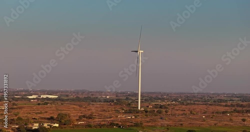 Golden hour aerial view of wind turbines across rural farmland, with rotating blades under a warm orange sky, showcasing clean energy and a serene countryside landscape of Gujrat, India