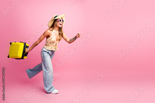 Fényképezés Young woman with yellow suitcase runs on pink studio background smiling in sunsh