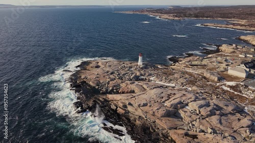 High drone aerial at sunset: historic Peggy's Cove lighthouse stands on snow-covered rocks against Atlantic. Halifax Nova Scotia Canada winter coastline, cinematic winter scene 4K.