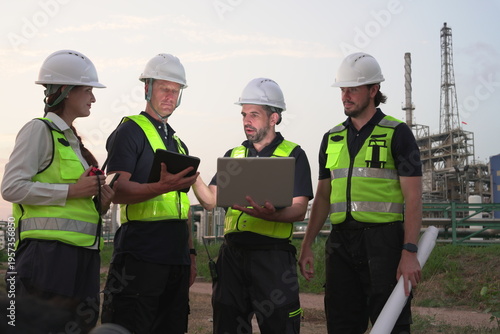 A group of male staff, male inspectors, female architects, male foremen, and managers, all wearing uniforms and helmets for safety, were reviewing blueprints, discussing plans at an oil refinery 