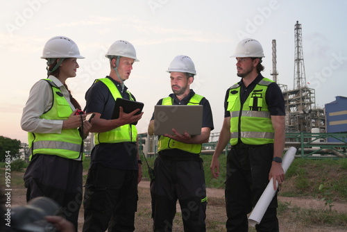 A group of male staff, male inspectors, female architects, male foremen, and managers, all wearing uniforms and helmets for safety, were reviewing blueprints, discussing plans at an oil refinery 