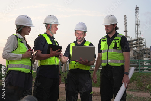 A group of male staff, male inspectors, female architects, male foremen, and managers, all wearing uniforms and helmets for safety, were reviewing blueprints, discussing plans at an oil refinery 
