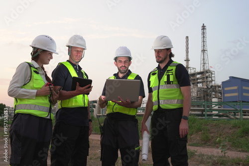 A group of male staff, male inspectors, female architects, male foremen, and managers, all wearing uniforms and helmets for safety, were reviewing blueprints, discussing plans at an oil refinery 