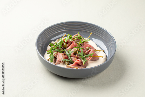 Thin sliced veal carpaccio with capers greens and creamy sauce served in gray bowl on light background