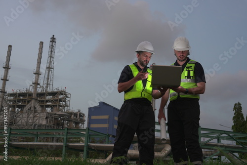 Male engineering staff and a male foreman/manager, all in uniform and helmets, are discussing and planning inspections of maintenance work at an oil refinery and petrochemical plant at night.

