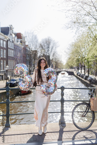 Smiling woman holding  33 balloons on a sunny Amsterdam canal bridge, spring birthday celebration