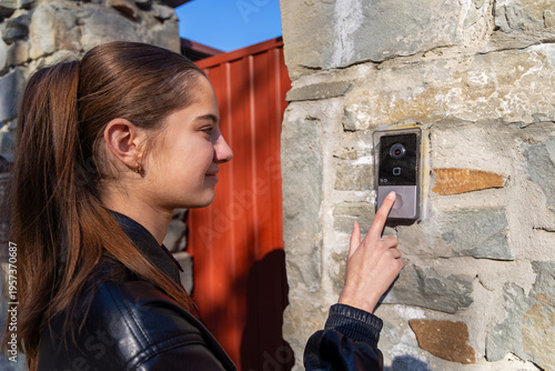 Young Woman Using Smart Doorbell at Gate. Side view of a young woman pressing a smart video doorbell on a stone gate, illustrating home security and visitor access.