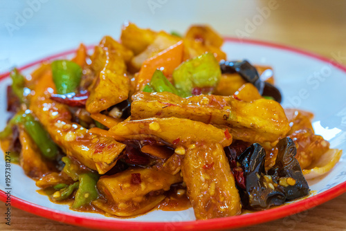 Close-up of tasty crispy eggplants in Chinese sweet and sour sauce with hot red chili pepper flakes on white plate. Chinese cuisine. Selective focus.