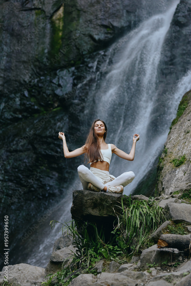 Naklejka premium Young woman meditating in white clothes sits cross-legged on rock near waterfall surrounded by lush greenery, expressing calmness and harmony in natural environment.