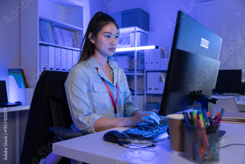 Asian woman working late night in modern office typing on keyboard looking focused at computer screen in blue ambient lighting showing dedication to corporate business project
