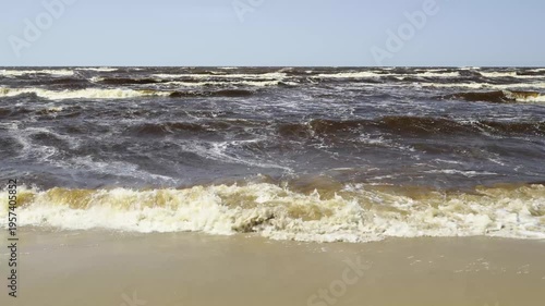 Sandy coastline of the Baltic Sea in Latvia. Yellow sand and large stones on the shore. Waves run onto the beach on a sunny day. Latvia, Gulf of Riga. 