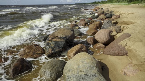 Sandy coastline of the Baltic Sea in Latvia. Yellow sand and large stones on the shore. Waves run onto the beach on a sunny day. Latvia, Gulf of Riga. 
