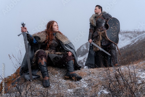 A fierce warrior woman with a sword rests on a rocky hill alongside a stoic knight amidst a snowy landscape.