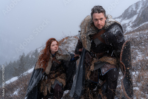 A snow-dusted couple, clad in winter furs and armor, gazes out over a snowy landscape.