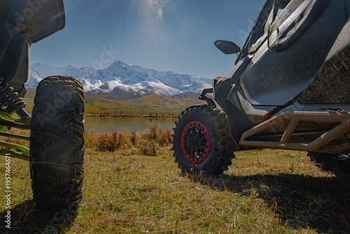 Close up of UTV off-road vehicles parked on a meadow by a mountain lake with snow peaks. Expedition, extreme adventure travel and overlanding concept.
