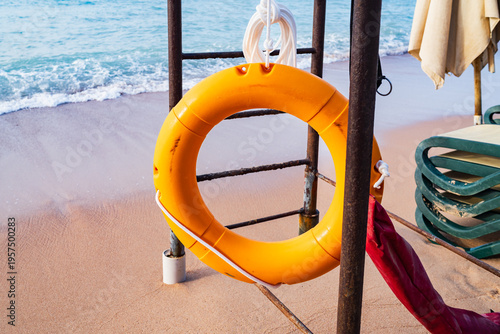 Bright Orange Lifebuoy Ring Hanging on a Lifeguard Station at a Sandy Tropical Beach with Blue Sea Waves in Background