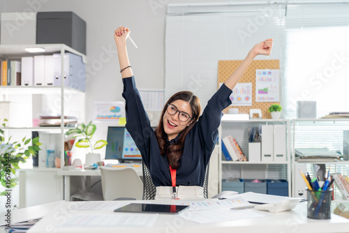 Successful Asian business woman stretching arms raised with joy sitting at office desk. Feeling happy after finish project achievement celebrate complete work career motivation productivity concept