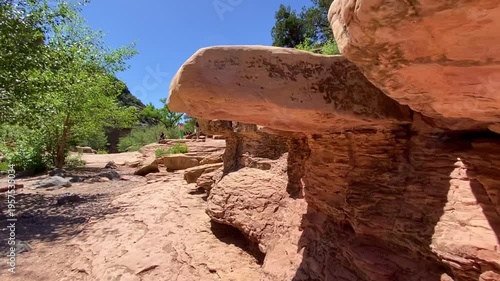 Smooth shot moving along massive red rock cliffs and hanging formations. Vibrant Arizona landscape with green trees, shrubs, and blue sky. Breathtaking canyon view at Slide Rock State Park.