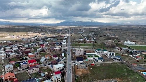 Aerial view of a quiet town nestled in a valley. Roads divide homes and green spaces. Cloudy sky looms above distant mountains. No people visible in this serene landscape