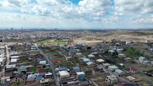 Aerial view shows a sprawling residential area under partly cloudy skies. Homes with varied rooftops stretch across rolling terrain. Green patches and roads divide neighborhoods