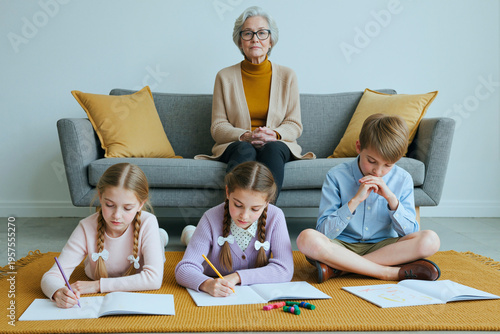Wide shot of senior woman supervising children drawing on floor at home, supporting caregiving and family learning