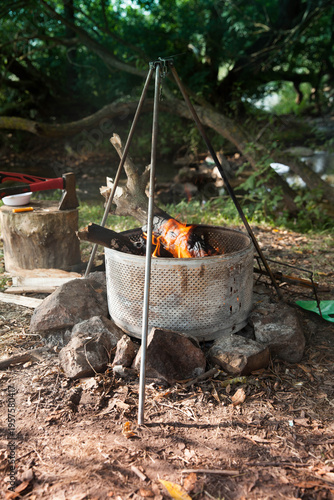 A fire burns inside a metal pot at a campsite surrounded by trees and stones during daylight hours