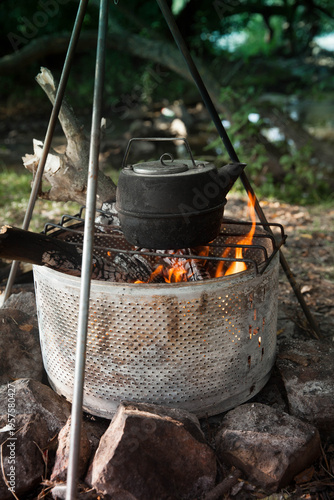 the kettle on a fire  at a campsite surrounded by trees and stones during daylight hours