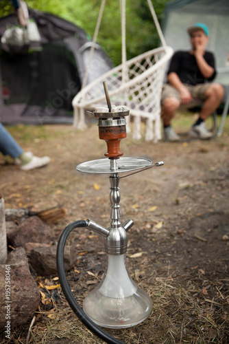 Friends enjoy hookah sitting near camping area with tents and swing in the background at summer evening