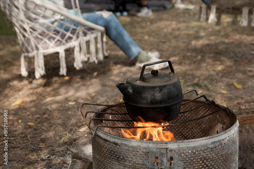 the kettle on a fire  at a campsite against the background of which a girl sits on a hanging chair during daylight hours