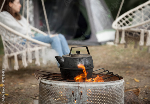 the kettle on a fire  at a campsite against the background of which a girl sits on a hanging chair during daylight hours