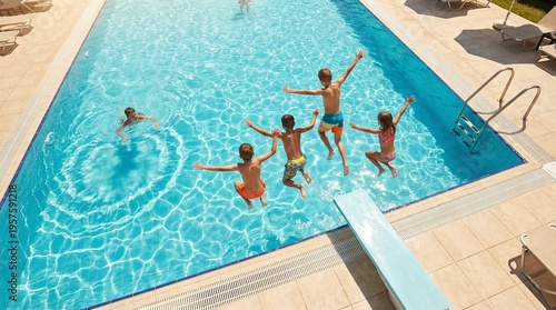 Group of happy children jumping into outdoor swimming pool from diving board on sunny summer day, fun vacation and leisure activity.