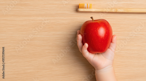 Red apple held by small hand with bamboo toothbrush on wooden surface, minimal style close up flat lay image showing healthy lifestyle and dental care