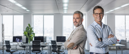 Two smiling confident business men leaders, happy corporate managers, young and middle aged professional executives looking at camera standing back to back with arms crossed in office. Portrait