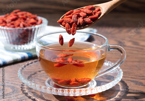 A wooden spoon with goji berries is tipping into a cup of tea, with a bowl of berries in the background