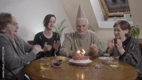 Senior man an party hat sitting at dinner table blowing candles on Birthday cake
