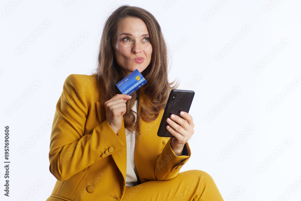 Naklejka premium A middle-aged businesswoman in a yellow suit holds a credit card and smartphone with a playful, upward glance, representing the ease and joy of modern digital finance.