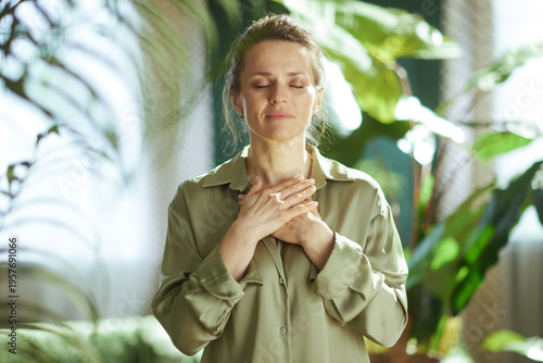 A middle-aged woman stands with her hands over her heart and eyes closed, engaging in a moment of self-compassion and emotional reset surrounded by greenery.