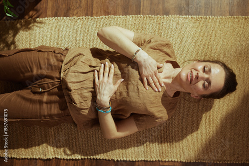 A woman in comfortable linen clothes lies on a woven mat, focusing on her breath to achieve inner peace and emotional balance during a home wellness ritual.