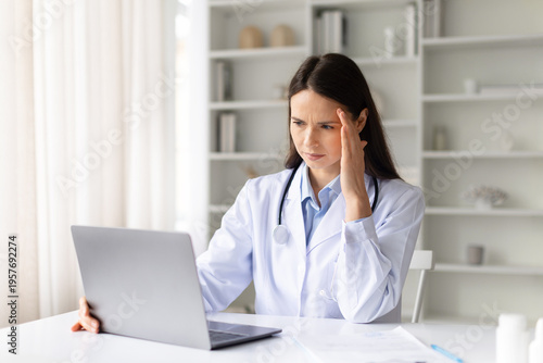 Slika na platnu Stressed female doctor working on laptop in clinic office, touching her temple while reviewing patient data