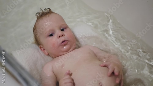 White baby lying in bathtub, gentle water, chubby cheeks and curious gaze, fingers exploring the skin of the tummy, towel supporting head, calm atmosphere capturing intimate bath routine, tender care