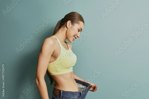 Woman smiles while holding oversized pants in front of green wall during indoor activity showcasing weight loss achievement
