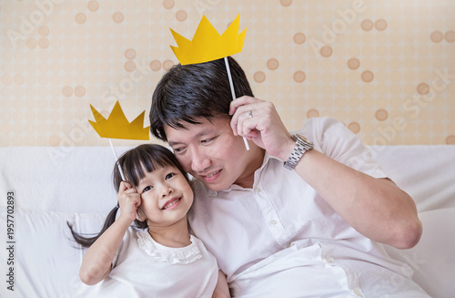 Billede på lærred Smile asian dad daughter lying on a bed together, holding yellow paper crowns and enjoying playful, quality time indoors