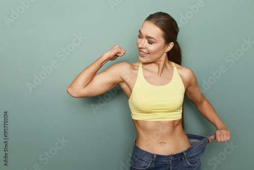 Woman shows off fitness progress while flexing arm muscles in casual outfit against a plain green background during daylight hours