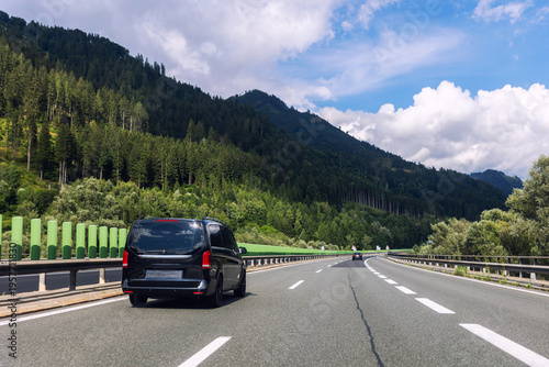 Luxury black minivan driving along alpine highway between green mountains blue sky. Comfort vip van transfer transportation passenger service, Airport shuttle taxi European mountain resorts