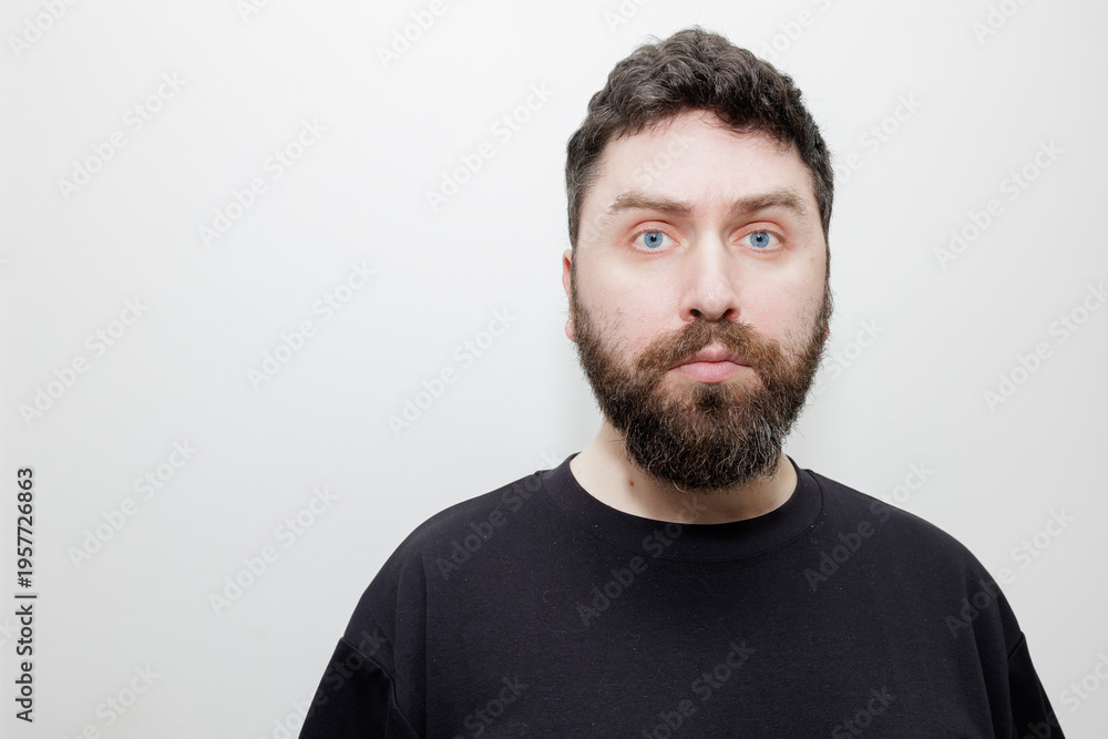 Fototapeta premium A direct, neutral headshot portrait of a man with dark hair, beard and blue eyes in a black t-shirt on a white background. Stock photo