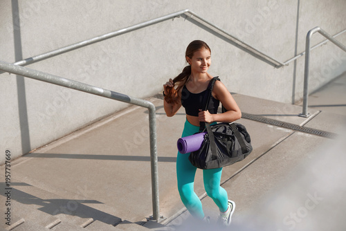 Young woman runs up the stairs carrying a fitness bag and yoga mat, dressed in trendy athletic clothing, showcasing a commitment to an active lifestyle.