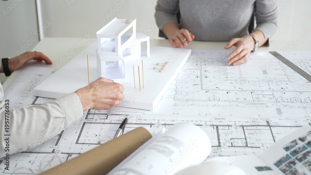 Obraz premium Architects collaborating, examining building blueprints with rulers and pens, developing innovative architectural designs and construction plans around a model house, close up