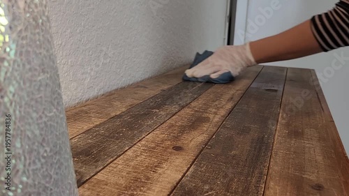 A woman wearing gloves wipes a wooden table with a cloth while moving a decorative object. Scene represents cleaning routine, hygiene, and home care.