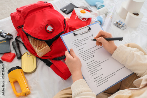 Young woman with checklist packing emergency backpack on bed, top view
